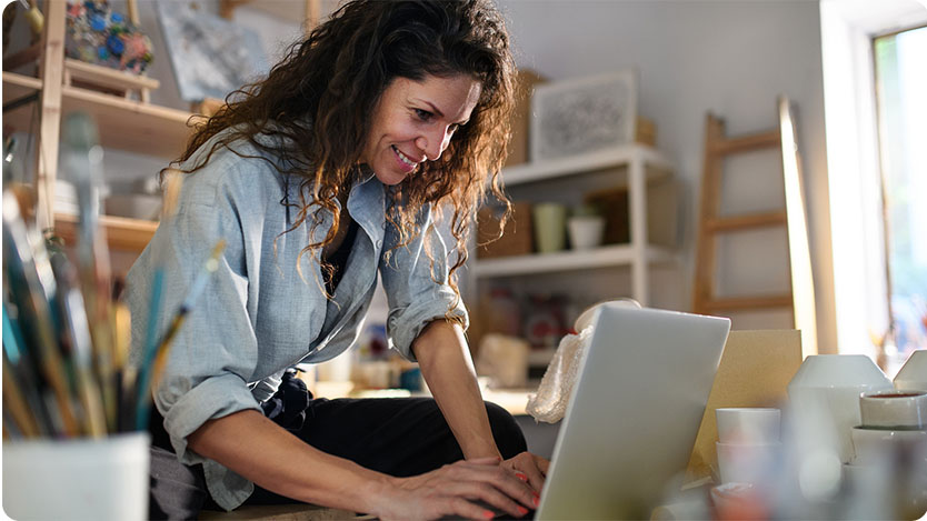 A person with long curly hair is working on a laptop in an art studio.
