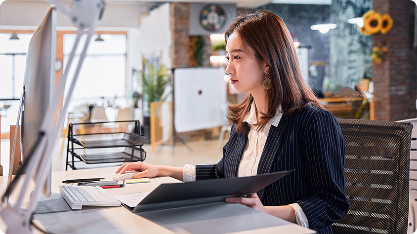 Person at an office desk holding a folder and looking at a large computer monitor in a modern workspace