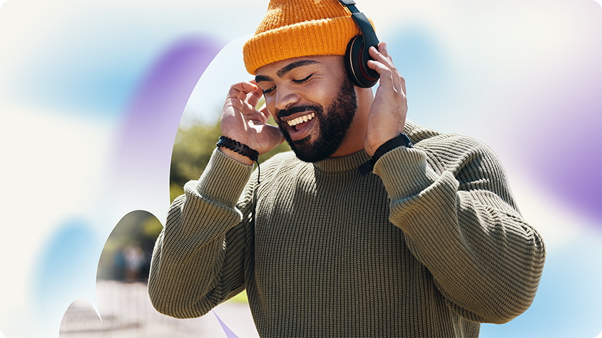 A person wearing a mustard‑yellow beanie and over-ear headphones adjusts the headphones while standing outdoors. They are dressed in a textured, olive-green sweater, with soft, colorful abstract shapes in the background.